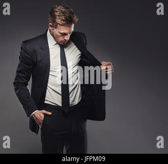 Suave giovane impreditore controllando la giacca di un elegante abito nero in piedi da solo in un monolocale contro uno sfondo grigio Foto Stock
