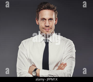 Bel giovane imprenditore che indossa una camicia e cravatta sorridente e in piedi con le braccia incrociate da soli in un studio contro uno sfondo grigio Foto Stock