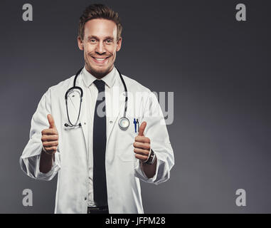 Sorridente giovane medico indossando un camice dando un entusiasta pollice alzato in piedi da solo in un monolocale contro uno sfondo grigio Foto Stock