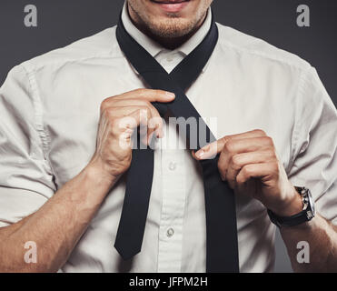 Primo piano di un giovane imprenditore in una camicia in piedi in un studio contro uno sfondo grigio la legatura la sua cravatta Foto Stock