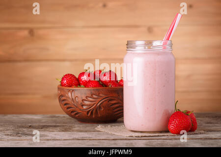 Frullato di fragole con paglia nel vasetto di vetro e fresche fragole mature nella ciotola vicino su sfondo di legno. Bibita salutare. Still Life Foto Stock