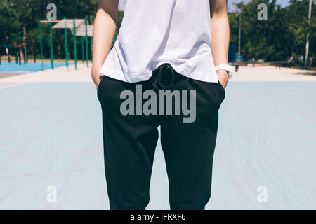 Immagine ritagliata della donna in pantaloni sportivi e t-shirt è in piedi con le mani nelle tasche sullo sfondo del campo sportivo, close-up Foto Stock