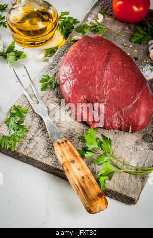 La cottura di filetto di manzo filetto. Grande tutto il pezzo di filetto di vitello su una vecchia scheda di taglio con una forcella per la carne e le spezie (pepe, sale, prezzemolo, aglio, Foto Stock