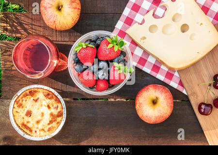 Foto aerea del picnic con mele, bicchiere di vino rosato, frutta fresca in contenitore di plastica, quiche, e il pezzo di formaggio Olandese, rustico di legno Foto Stock