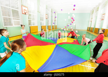 Ritratto di felice preteen i ragazzi e le ragazze in piedi in cerchio, salutando paracadute arcobaleno pieno di palline colorate, in sports hall Foto Stock