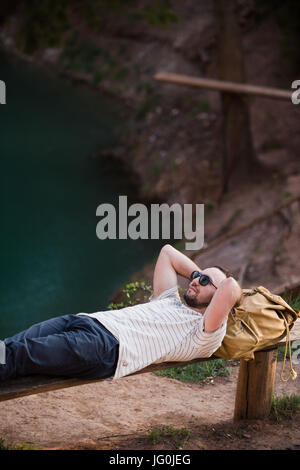 L'uomo rilassante all'esterno. Indossando un bianco di T-shirt, pantaloni scuri, occhiali da sole e un giovane ragazzo è sdraiato sul banco di legno contro il lago in una foresta all'aperto Foto Stock