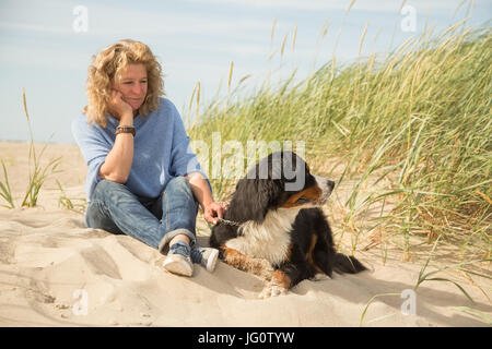 Donna matura con il suo cane sulla sabbia e erba seduta Foto Stock