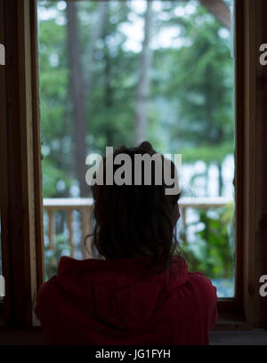 Bruna giovane femmina dai capelli a guardare fuori dalla finestra di casa al bosco in un giorno di pioggia. Foto Stock