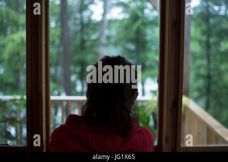 Bruna giovane femmina dai capelli a guardare fuori dalla finestra di casa al bosco in un giorno di pioggia. Foto Stock