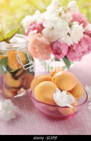 Giallo maturo le albicocche in un vaso trasparente con un detox bevanda fatta di menta e albicocche secche in background con bouquet di peonie. Foto Stock