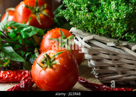 Organici freschi pomodori maturi con gocce d'acqua, il timo in cesto in vimini, verde e viola, basilico, peperoncino su legno tavolo da cucina, rustico Provence, me Foto Stock