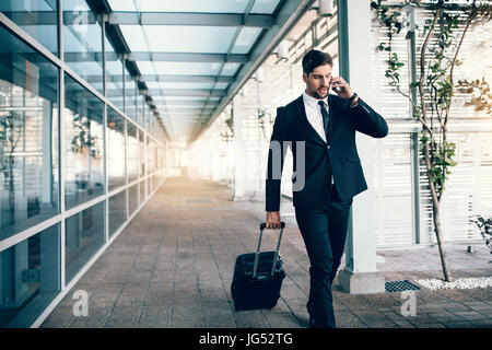 Bel giovane uomo in viaggio d'affari a piedi con il suo bagaglio e parlando al cellulare all'aeroporto. Viaggia per lavoro effettuare chiamate telefoniche. Foto Stock