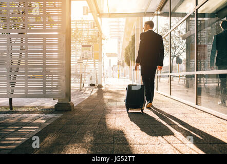 Vista posteriore di imprenditore a piedi con il sacchetto al di fuori dell'aeroporto. Giovani viaggiatori di affari tirando la valigia sulla strada della citta'. Foto Stock