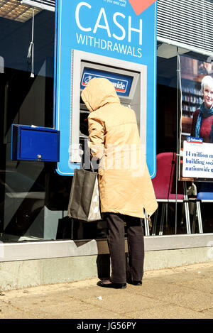 Donna anziana con un cappotto con cappuccio utilizza un ATM cash point machine di un Nationwide building society bank in Essex, Inghilterra Foto Stock