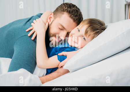 Padre abbracciando sorridendo piccolo figlio giacente in letto di ospedale, papà e figlio in ospedale Foto Stock