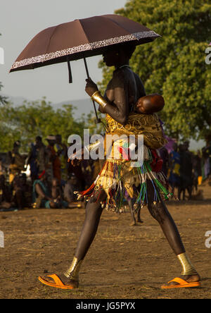 La donna che porta il suo bambino sulla schiena con un ombrello durante il grasso uomini cerimonia in Bodi tribù, valle dell'Omo, Hana Mursi, Etiopia Foto Stock