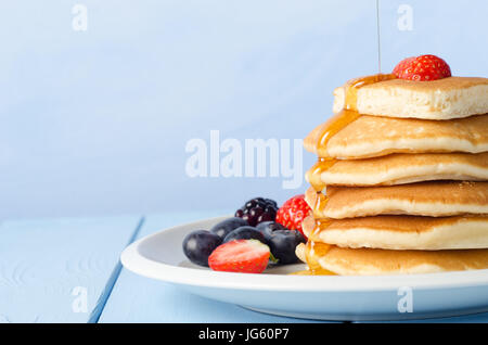 Una pila di frittelle di colazione e rabboccato con una fragola, in piedi su un bianco piastra Cina, circondato da frutta estiva, con sciroppo d'acero versata fr Foto Stock