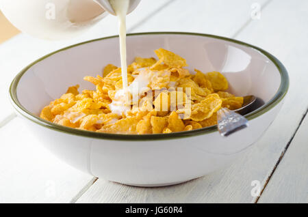 Versare il latte da una brocca in una ciotola di cornflake colazione, su un dipinto di bianco planked legno tavolo da cucina. Foto Stock