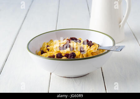 Una prima colazione in scena con una verde bordato bianco di ceramica ciotola di cornflake, uvetta e cucchiaio con un bricco per il latte in soft focus dietro di esso. La tabella è usurato vecchio Foto Stock