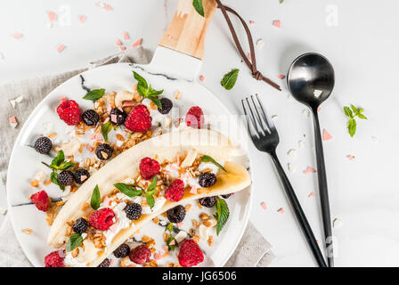 Frutta estiva berry prima colazione. Una sana banana split colazione con crema di formaggio, lamponi, more, menta, bianco e rosa di cioccolato. Su bianco ma Foto Stock