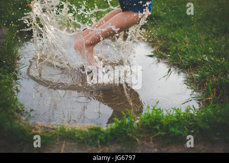 Un bambino salta in una pozza di fango con spruzzi di acqua attorno ai suoi piedi. Foto Stock