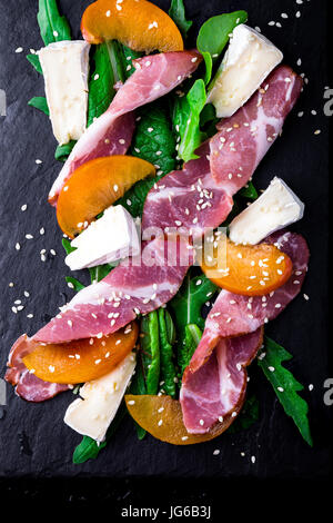 Insalata con prosciutto jamon serrano, camembert, melone, rucola su nero ardesia pietra piastra su sfondo nero. Vista dall'alto. Foto Stock