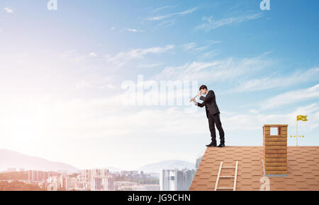 Giovane imprenditore determinato in piedi sul tetto di casa e guardando a cannocchiale. I supporti misti Foto Stock