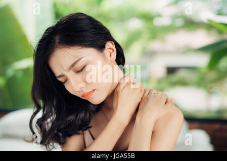 Giovane donna asiatica che soffrono di dolore al collo mentre è seduto sul letto di casa Foto Stock