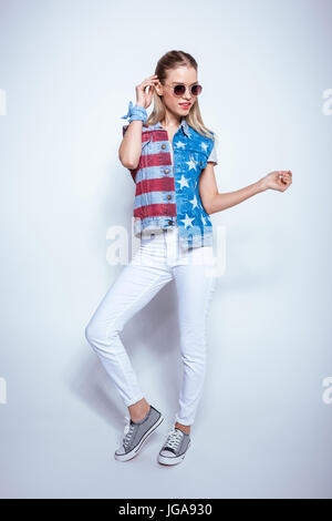 Attraente ragazza bionda in occhiali da sole indossando giubbotto in denim con noi bandiera e in posa di studio Foto Stock