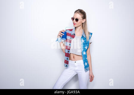 Ragazza sorridente in occhiali da sole indossando giubbotto in denim con stelle e strisce e bere soda da alluminio può Foto Stock