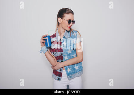 Tanga elegante ragazza bionda americana outfit patriottica e occhiali da sole tenendo il carbonato di sodio può isolati su grigio, cultura americana concept Foto Stock