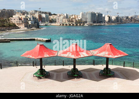 Tre ombrelli rosso o ombrelloni su panchine pubbliche sul lungomare a Balluta Bay a St Julians Malta su una soleggiata giornata estiva Foto Stock