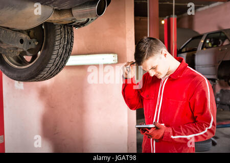 Giovane maschio confuso motoriste rendendo permanente note sotto un sollevato in auto nei pressi di attrezzatura di lavoro per la riparazione e di diagnosi nella riparazione auto servi Foto Stock