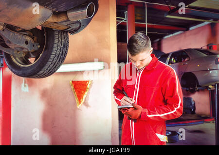 Giovane maschio meccanico del motore rendendo permanente note sotto un sollevato in auto nei pressi di attrezzatura di lavoro per la riparazione e di diagnosi automatica del servizio di riparazione Foto Stock