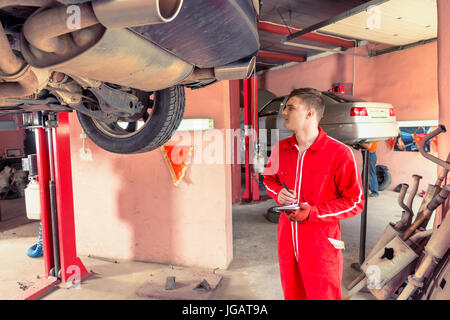 Bel maschio meccanico del motore rendendo permanente note sotto un sollevato in auto nei pressi di attrezzatura di lavoro per la riparazione e di diagnosi automatica del servizio di riparazione Foto Stock