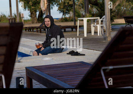 Studente in una felpa con cappuccio a bere birra e gambe di ribaltamento in acqua di piscina Foto Stock