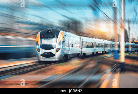 Elevata velocità del treno per pendolari presso la stazione ferroviaria al tramonto in Europa. Moderno treno intercity sulla piattaforma ferroviaria. Vista urbano con belle passeggero Foto Stock