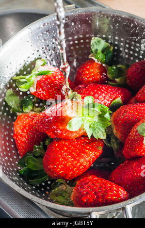 Un vaglio metallico in appoggio in un lavello da cucina, riempito con fragole fresche e di essere lavato sotto l'acqua corrente. Foto Stock