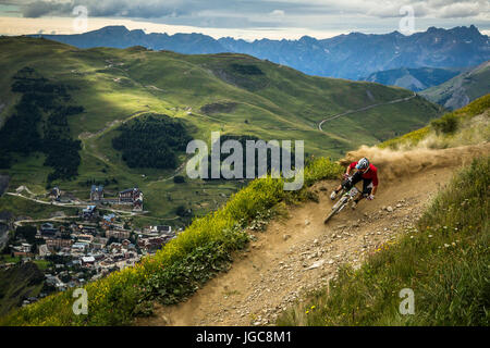Un mountain biker cavalca un angolo polveroso alta nelle alpi. Il villaggio di sci di Les Deux Alpes in background Foto Stock