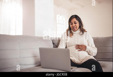 Ritratto di giovane e bella donna incinta con il computer portatile seduti sul divano. Donna africana in attesa di un bambino seduto sul divano e utilizzo portatile a casa. Foto Stock