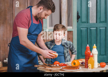 Carino piccolo ragazzo con il padre nel grembiule preparazione di hot dog insieme nel cortile, papà e figlio concetto di cucina a vista Foto Stock