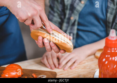 Close-up vista parziale del ragazzino e padre bun di taglio per hot dog, papà e figlio concetto di cucina a vista Foto Stock