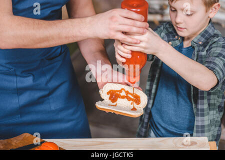 Ritagliato colpo di padre e figlio versando la salsa in hot dog, papà e figlio concetto di cucina a vista Foto Stock