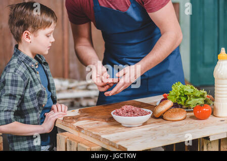 Padre nel grembiule e carino piccolo figlio di preparare hamburger insieme, papà e figlio concetto di cucina a vista Foto Stock