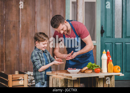 Padre nel grembiule e carino piccolo figlio di preparare hamburger insieme, papà e figlio concetto di cucina a vista Foto Stock