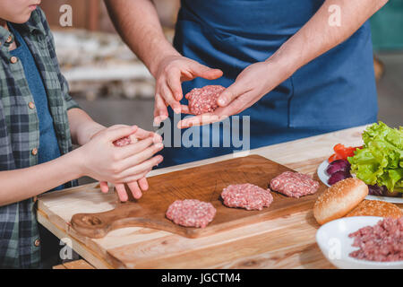 Close-up vista parziale del figlio e del padre la cottura di hamburger sul tagliere di legno, papà e figlio concetto di cucina a vista Foto Stock