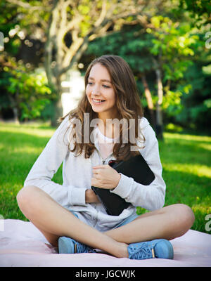Donna sorridente con bretelle seduti in giardino su una coperta tenendo la sua tavoletta digitale Foto Stock