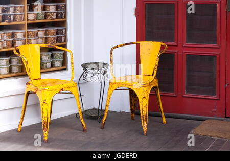 Due colorato giallo weathered sedie in metallo su un portico addossato a una porta rossa. Foto Stock