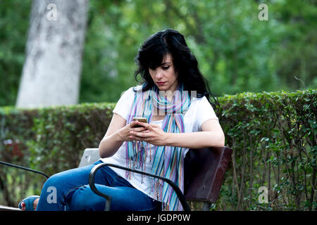 Attraente bella latino donna seduta su una panchina nel parco sensazione triste e depresso sul suo smart phone in un parco Foto Stock