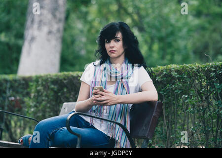 Attraente bella latino donna seduta su una panchina nel parco sensazione triste e depresso sul suo smart phone in un parco Foto Stock